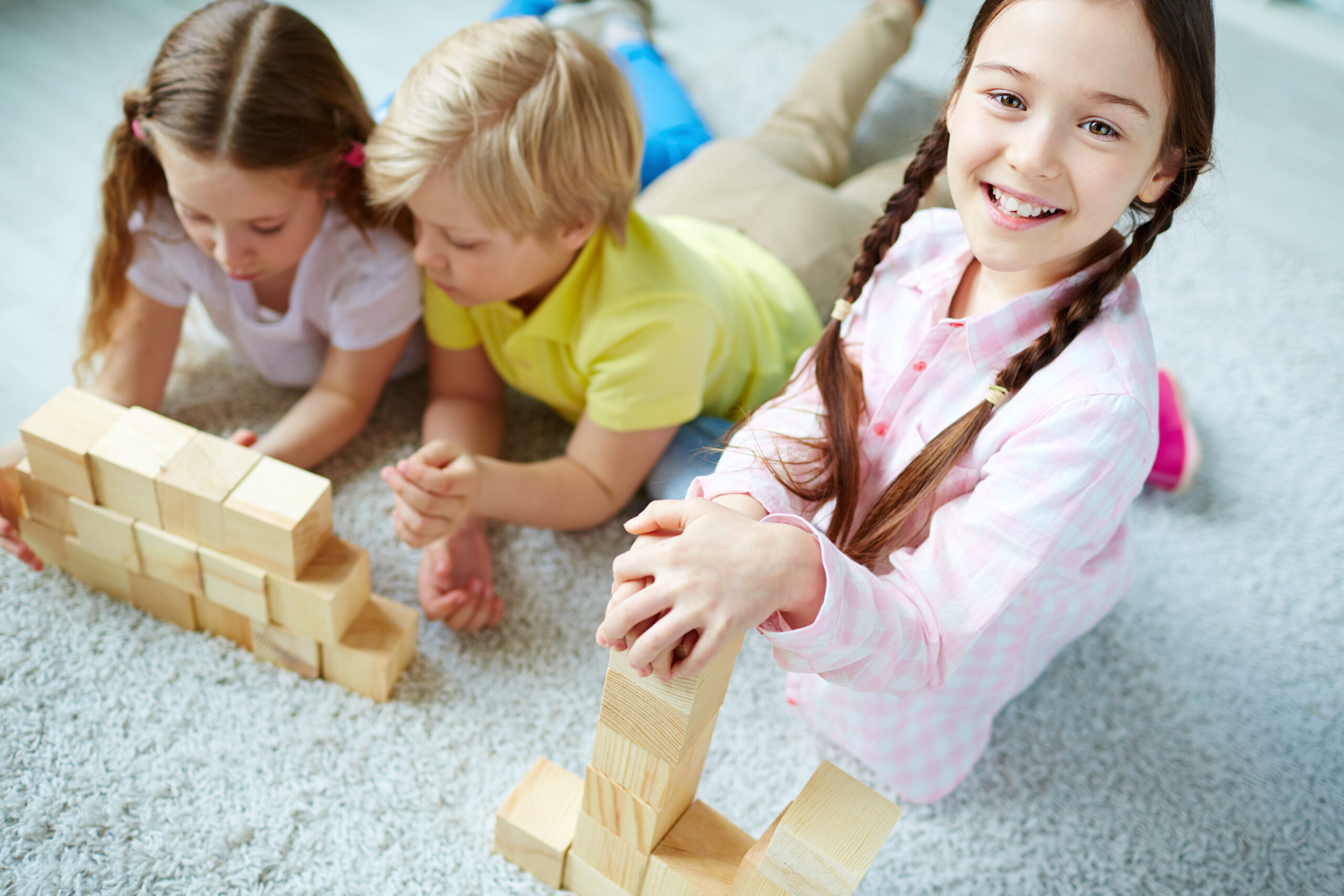 Three little friends playing with wooden bricks, happy girl looking at camera